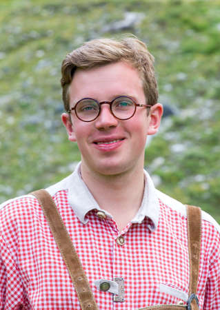Beautiful portrait of a handsome young man with glasses in traditional clothing with lederhosen and checkered shirt in summer in the alps near Grimentz, Switzerlandの写真素材