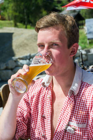 Beautiful portrait of a handsome young man in traditional clothing with checkered shirt enjoying a cold beer in summer in Grimentz, Switzerlandの写真素材