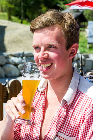 Beautiful portrait of a handsome young man in traditional clothing with checkered shirt enjoying a cold beer in summer in Grimentz, Switzerlandの写真素材