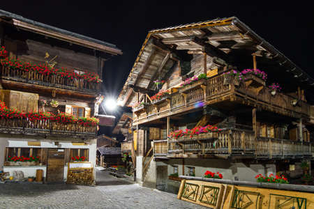 Beautiful traditional wooden houses in the streets of the alpine village Grimentz, Switzerland, canton Valais, municipality Anniviers, at night with geranium flowers on the balconies illuminated by streetlightsの写真素材