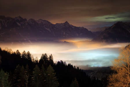 Beautiful winter night view of the city Brand in the valley of the Brandnertal in the mountains of the Alps in Vorarlberg, Austria, with illuminated fog cloudsの写真素材