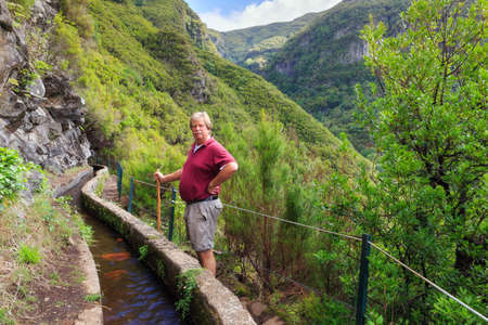 Beautiful landscape view of a senior tourist on the hiking path in nature on the green island Madeira, during a hike on a trail along a famous levadaの写真素材