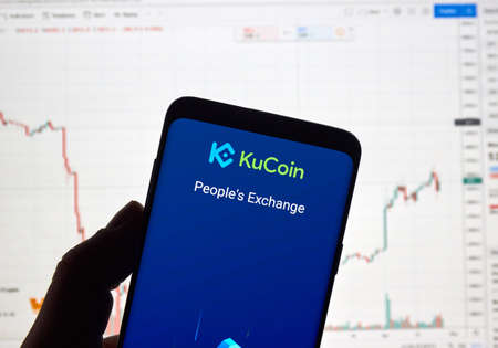 MONTREAL, CANADA - APRIL 26, 2019: Kucoin, cryptocurrency exchange logo and application on Android Samsung Galaxy s9 Plus screen in a hand over a laptop display with bitcoin chart on it.のeditorial素材