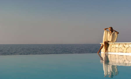 Greek style woman sitting on edge of infinity pool at sunsetの写真素材