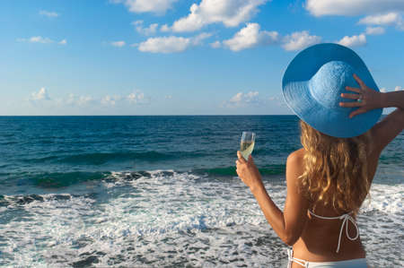 Sexy woman in bikini and hat with glass of wine looking at the sea. Crete. Greece の写真素材