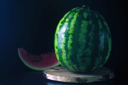 Ripe whole watermelon stands on a wooden board against a black backgroundの写真素材