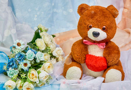 Teddy bear with a red heart in hands and a bouquet of flowers on a colored festive background with place for text.Copy spaceの写真素材