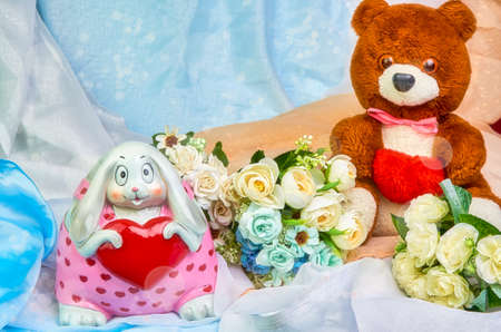 A teddy bear with a red heart in his hands, and a bunny with a heart in his hands, a bouquet of flowers on a colored holiday background with place for text. Valentine's Day. Postcardの写真素材