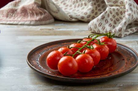 Cherry tomatoes on a green branch lie in a brown clay plate with drops of water. Close-up.の写真素材
