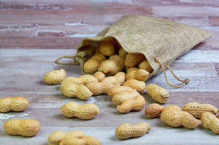 Inshell peanuts fell out of a linen bag against a wooden background. Horizontally close up.