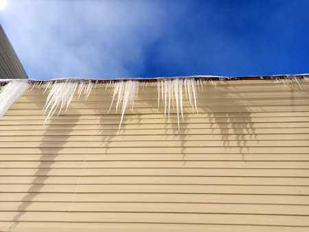 icicles hanging from the roof against a blue sky and a beige wallの写真素材