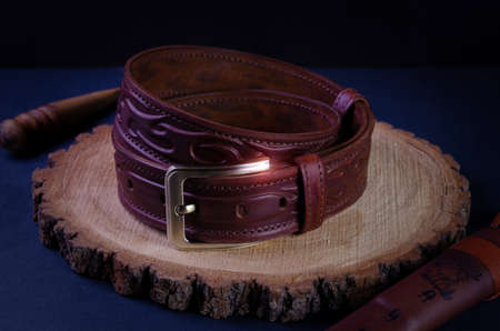 A brown, men's handmade belt made of genuine leather, sewn with a thread with wax and a brass buckle on a dark background, lies on a tree saw cut.の写真素材