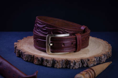A brown, men's handmade belt made of genuine leather, sewn with a thread with wax and a brass buckle on a dark background, lies on a tree saw cut.の写真素材