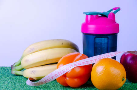 Measured blue bottle with a pink cap, a shaker for sports nutrition on green grass, measuring tape with bananas, red apple, orange bell pepper, orange on a white background, diet.Copy space.の写真素材
