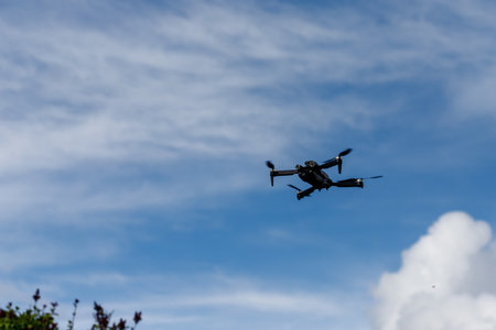 A black drone hovers in the air against a backdrop of blue sky with scattered white clouds. Some vegetation is visible in the bottom left corner.の写真素材