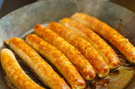 Close-up of several browned sausages sizzling in a pan, with visible steam rising and a golden-brown crust.の写真素材