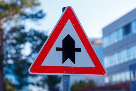 A triangular warning sign with a red border displays a black symbol representing a priority road merging with a side road.の写真素材