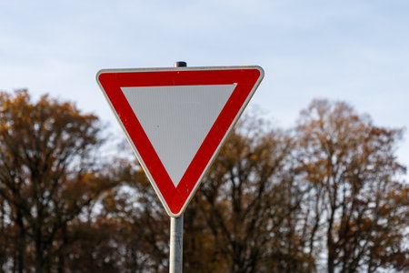A triangular traffic sign with a red border and a white center is mounted on a metal pole, set against a blurry background of autumn trees and a pale sky.の写真素材