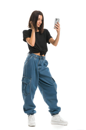 A young woman is taking a selfie with her phone while wearing a black shirt and blue denim cargo jeans, suitable for a casual fashion look.の素材