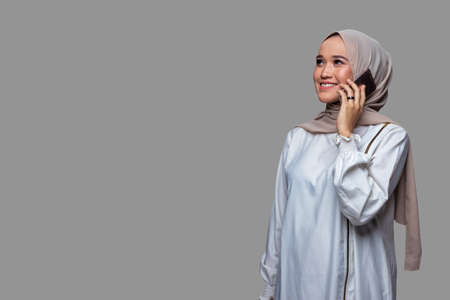 Beautiful woman wearing hijab is making a phone call with a smiling expression while looking up with plain spaceの写真素材