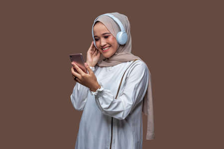 Woman in hijab wearing wireless headphones while looking at the cellphone screenの写真素材