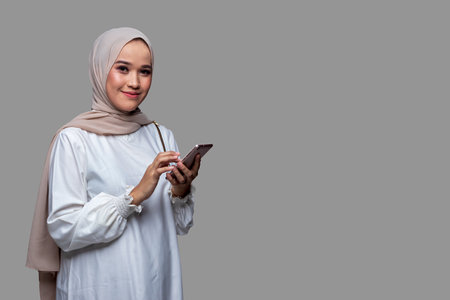 Beautiful woman in hijab is holding a cellphone smiling and looking at the camera isolated on plain backgroundの写真素材