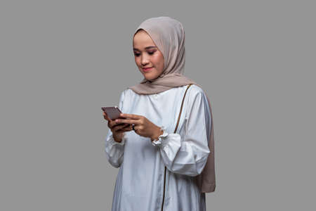 Beautiful hijab woman is typing a message on her cell phone with smile expression isolated on plain backgroundの写真素材