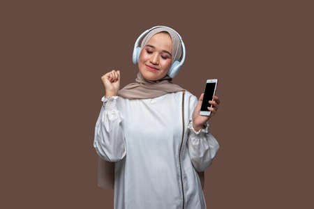 Woman in hijab is listening music from her phone using wireless headphones while dancing and closing her eyesの写真素材