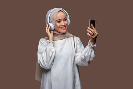 Asian woman in hijab wearing wireless headphones while looking at the cellphone screenの写真素材