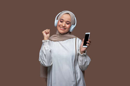 Young woman in hijab is listening music from cell phone using wireless headphones while dancing and closing her eyesの写真素材