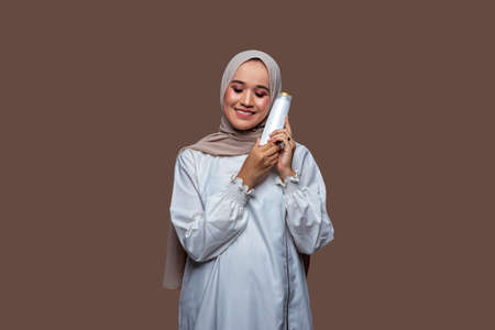 Beautiful woman in hijab is holding a shampoo bottle with both hands and putting it on her left cheek while closing her eyes and smilingの写真素材