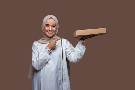 Hijab woman holding a pizza box with one left hand and her right hand pointing while smilingの写真素材