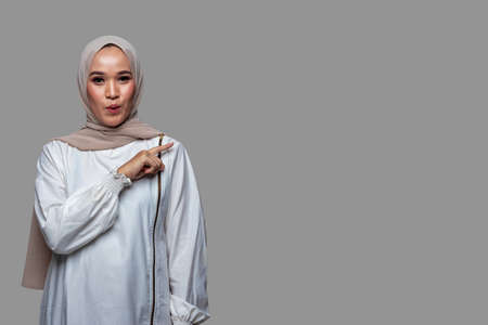 Beautiful muslim woman in hijab is pointing to the right side with shocked expression isolated on plain backgroundの写真素材