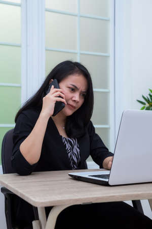 Woman sitting on chair with laptop at table holding drinking glass and looking at screenの写真素材