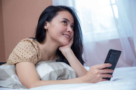 Beautiful woman is imagining on the bed while holding a cell phoneの写真素材