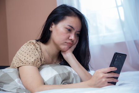 Woman lying on bed with duck lips expression while holding cell phoneの写真素材