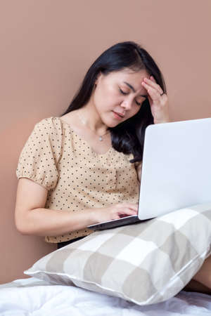 Woman with laptop on bed with hands holding forehead while closing her eyesの写真素材