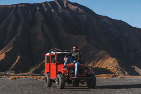 Cool guy sitting on red offroad jeep with Bromo mountain background. Tourist visiting Semeru...
