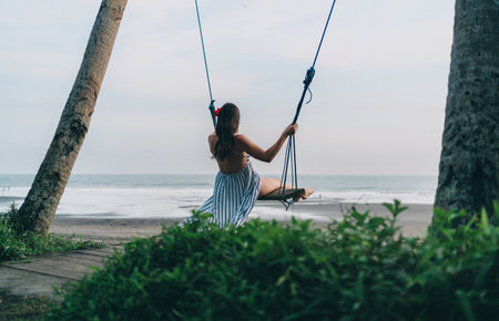 Back view of girl swinging on a swing with wonderful sea view. Young lady on tropical beach swingの写真素材