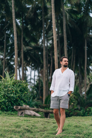 Portrait of casual dressed young man with jungle palms background. Guy in white shirt and shorts enjoy tropical nature environmentの写真素材