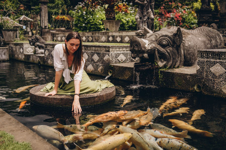 Beautiful girl sitting and admiring pond koi fishes. Smiling girl feeding carps in Ubud Water Palaceの写真素材
