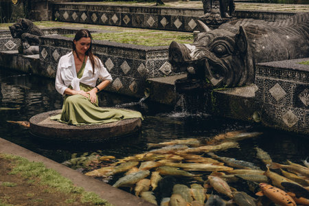 Beautiful girl sitting and admiring pond koi fishes. Smiling girl feeding carps in Ubud Water Palaceの写真素材
