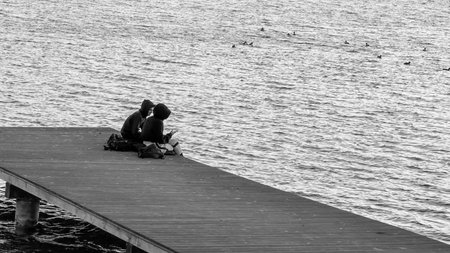 Two people sitting on a pier by the water, enjoying a peaceful moment with ducks swimming nearby, capturing tranquility and nature.の写真素材