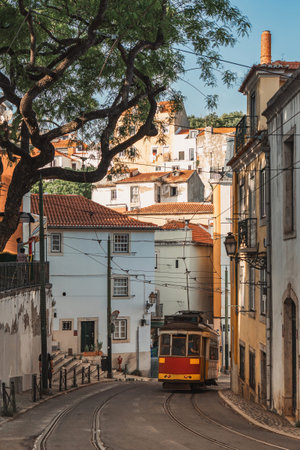 Charming yellow tram travels through narrow, winding streets of Lisbon Alfama district, surrounded...