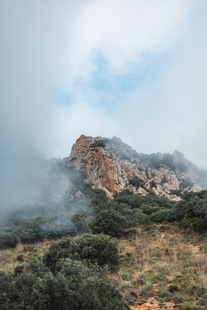 Dramatic rocky mountain peak shrouded in mist and clouds, lush green vegetation, and rugged terrain in the scenic Puig Campana region, Spain. Perfect for nature, travel, and adventure themes.の写真素材