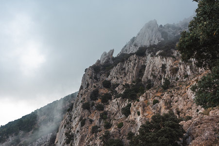 Steep rocky cliffs of Puig Campana in Spain rise through mist and clouds, creating a dramatic and moody mountain landscape ideal for nature, travel, and adventure themes.の写真素材