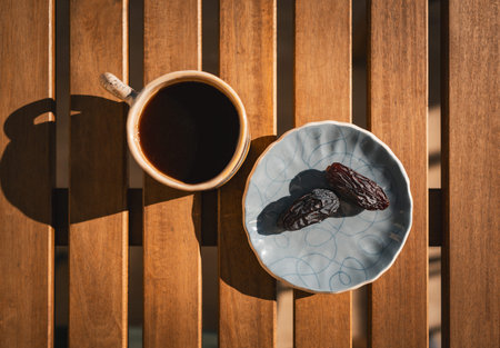 Top view of a cup of black coffee and two dried dates on a ceramic plate, arranged on a wooden table...