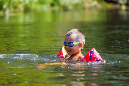 Cute boy is swimming in the small river with his armbands and gogglesの写真素材