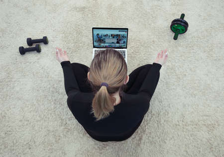 Rear view, Athletic healthy woman sitting on mat in lotus position, wearing sports black uniform, watching online yoga classes, doing breathing exercises on laptop. sports equipment is nearby.の写真素材