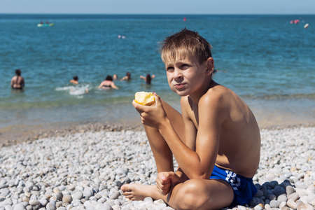 a tanned blond boy with blond hair is sitting on the beach eating an apple near the sea in blue shortsの写真素材